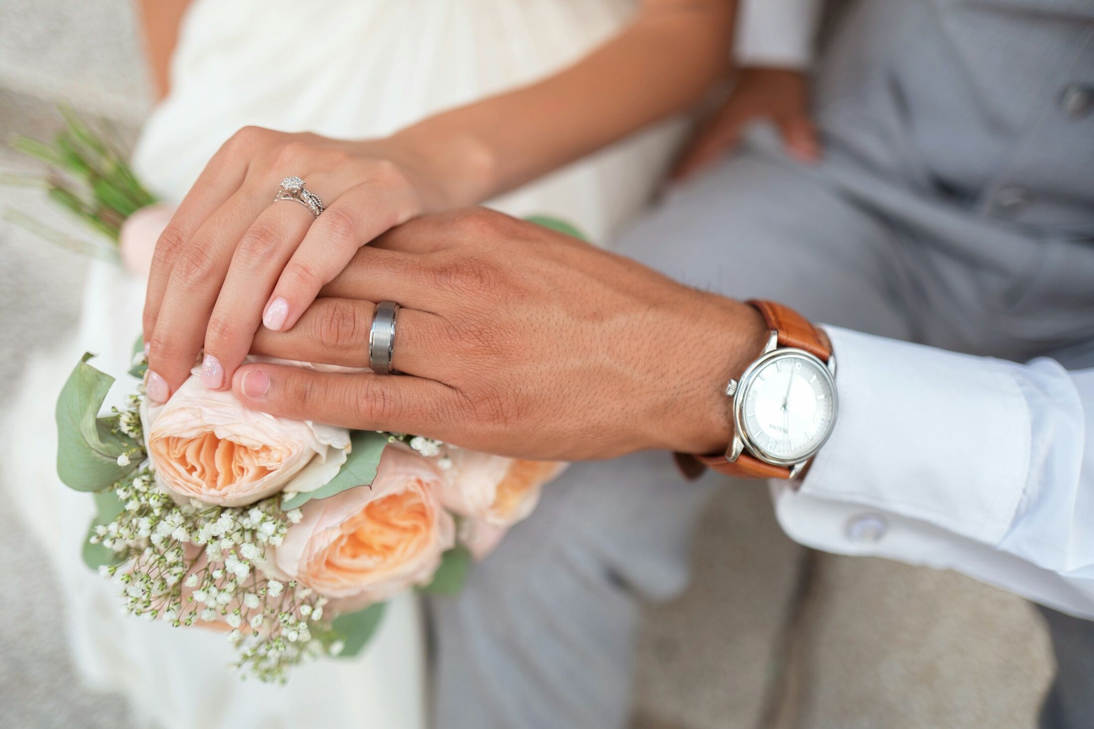 A couple holding hands, wedding rings and a bouquet of peach roses